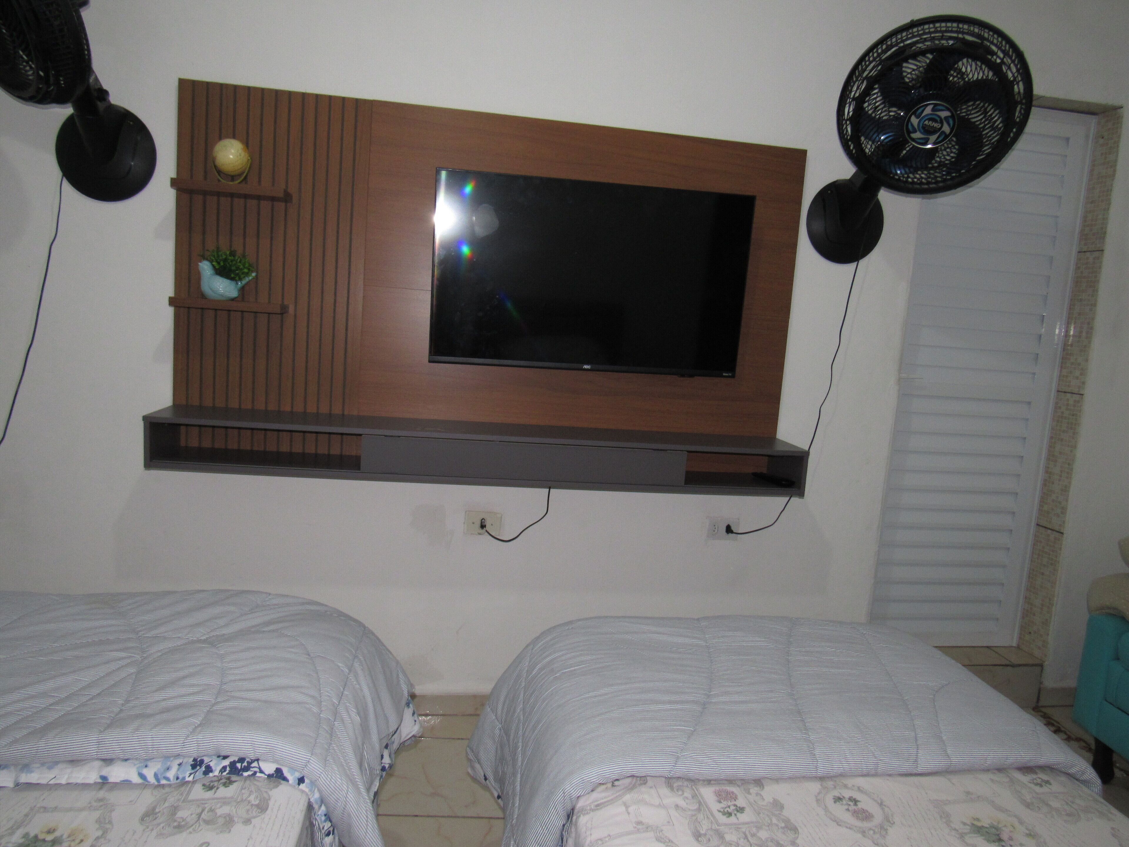 Ein Schlafzimmer mit an der Wand montiertem Fernseher, zwei Betten mit Blumenmuster auf der Bettwäsche und einer kleinen Ablage mit Dekoration.