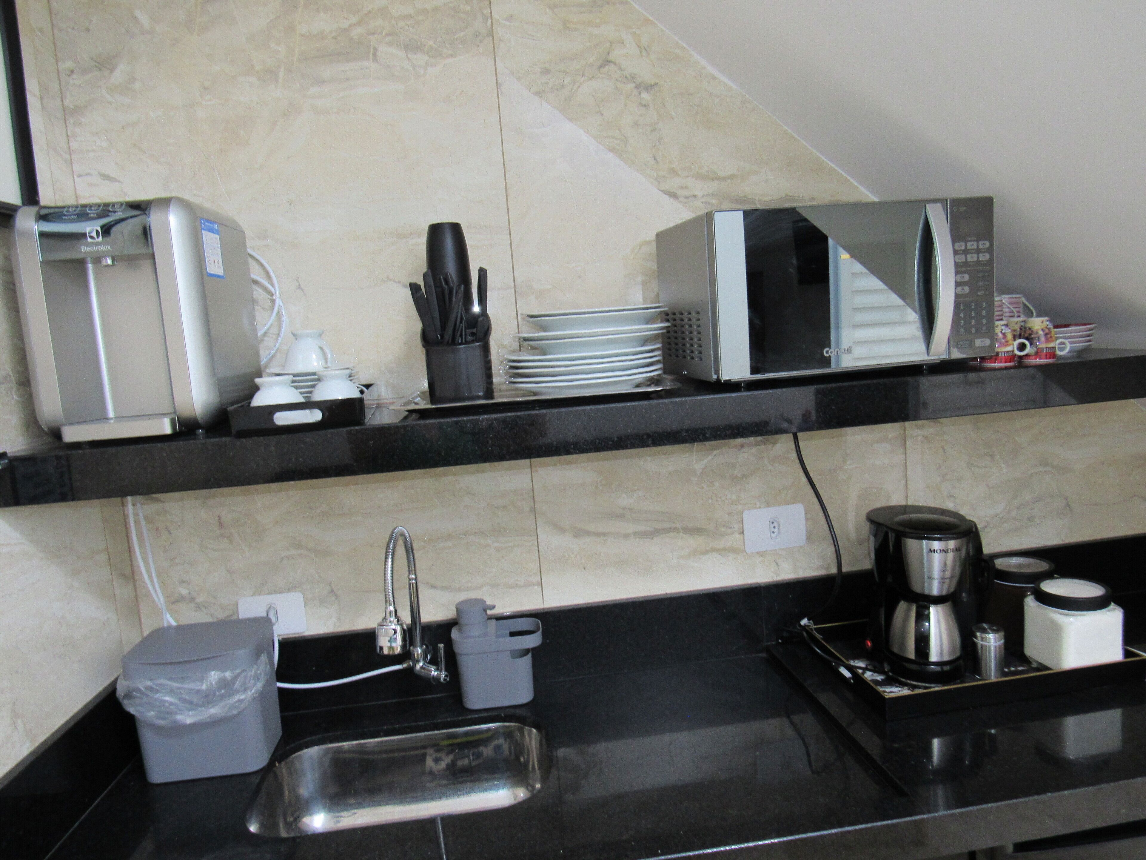 Cozinha equipada com micro-ondas, torradeira e máquina de café. Há uma pia com torneira e um cesto de lixo.