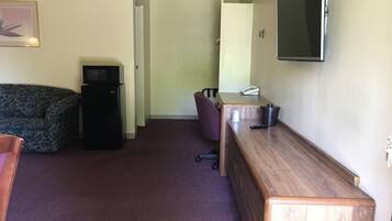 Habitación de hotel compacta con sofá de color oscuro, un refrigerador pequeño negro, un escritorio de madera con teléfono y control remoto, y un televisor montado en la pared.
