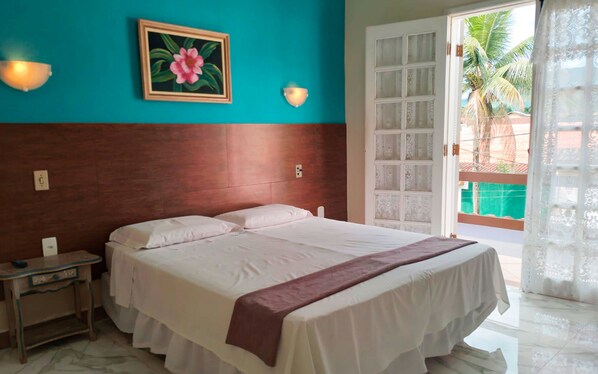 A hotel room with a bed, a nightstand, a painting, and a view of the outside. - Pousada Larissa (Paraty)