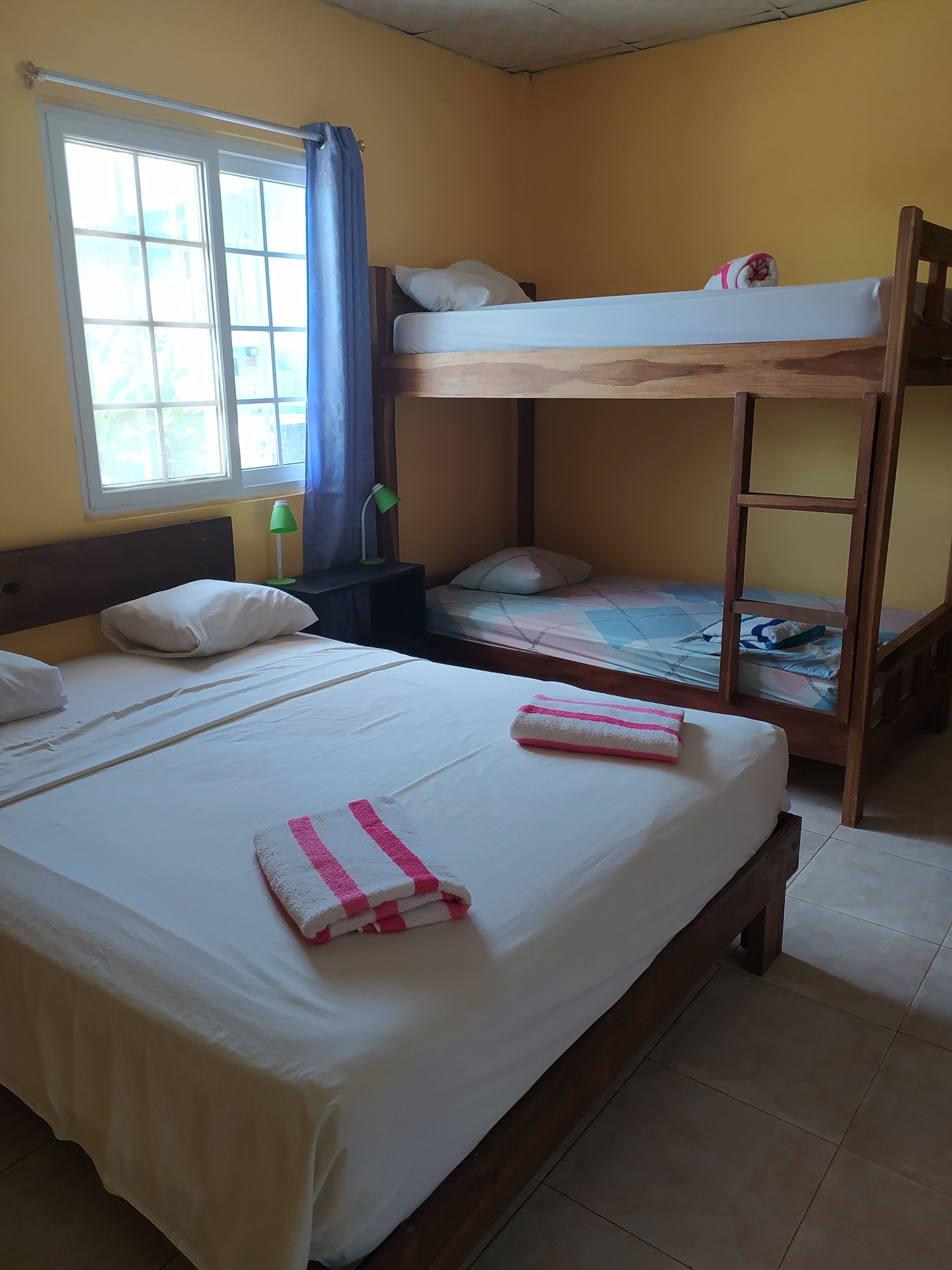 Una habitación con una cama litera, una cama individual, una mesita de noche, una lámpara y una ventana con cortinas.