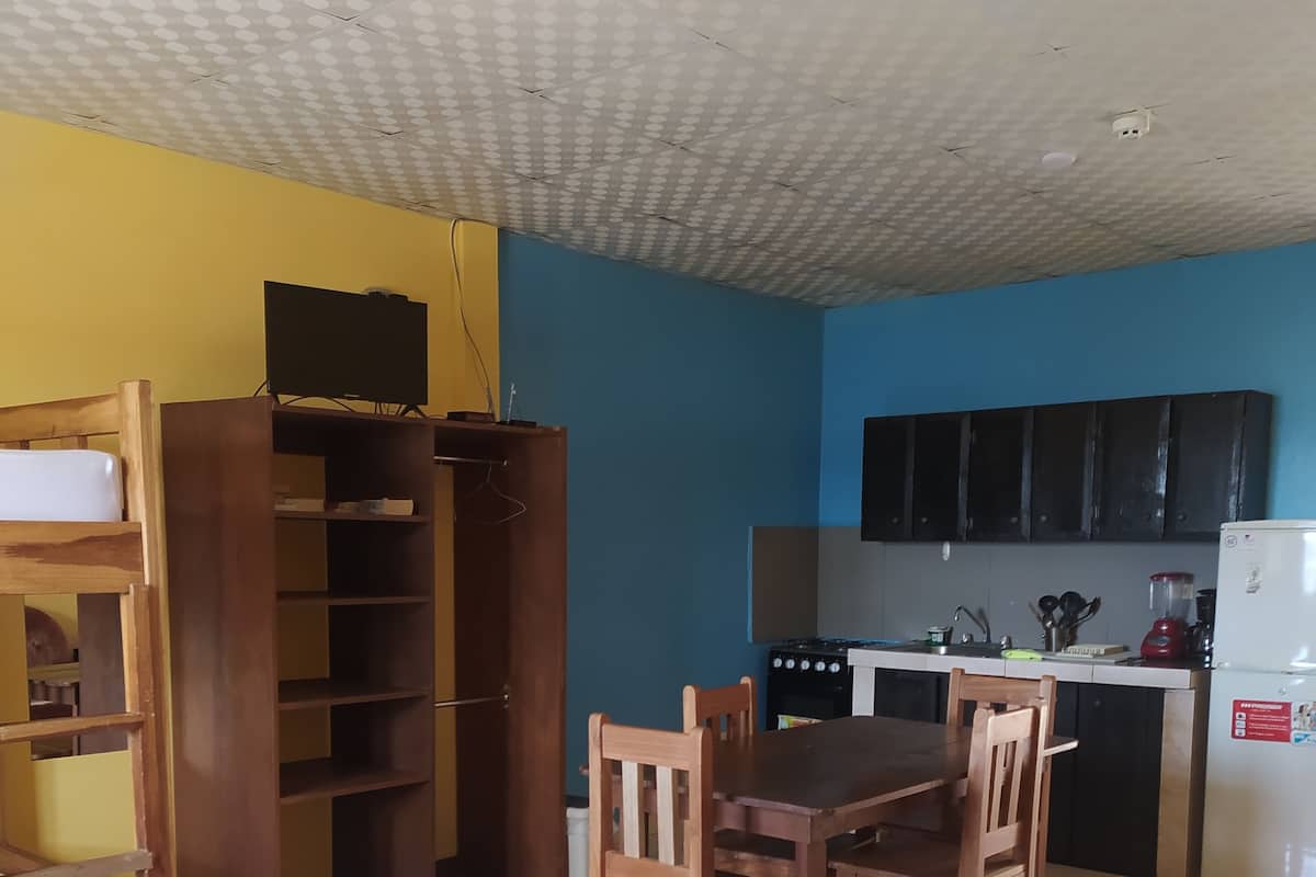 A room with a bed, a wooden desk, a dining table with chairs, a kitchen area with cabinets and appliances, and a ceiling with a grid pattern.