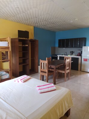 Een kamer met een bed, een houten bureau, een eettafel met stoelen, een keuken met kasten en apparatuur, en een plafond met een rasterpatroon.