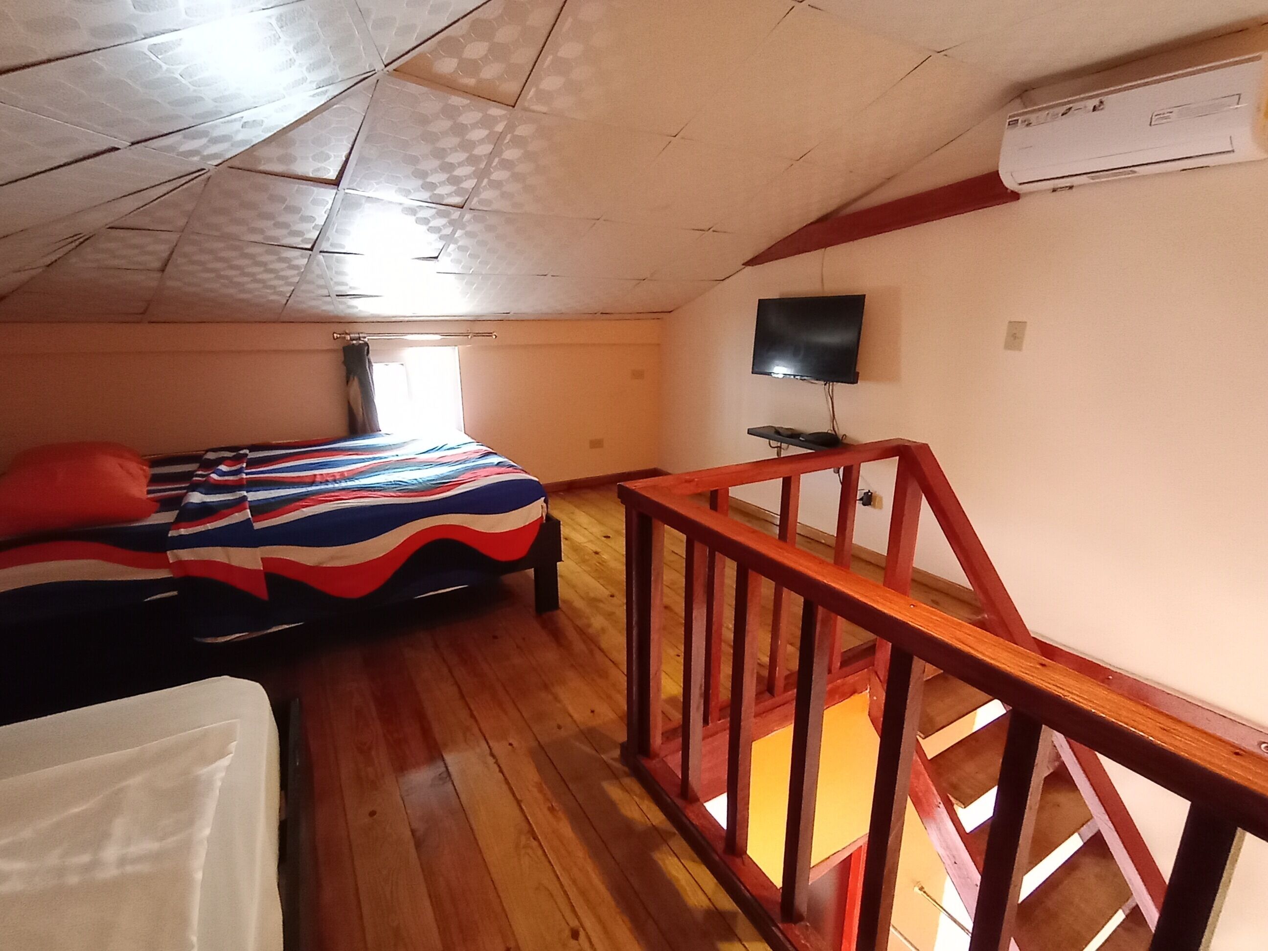 A bedroom with a bed, a wooden floor, a TV mounted on the wall, and a window with curtains.