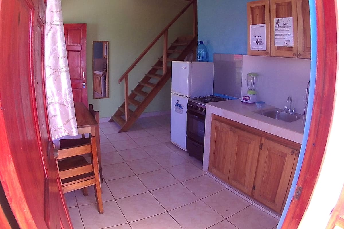 A compact kitchen with a refrigerator, stove, and sink. A wooden chair and a staircase are also visible.