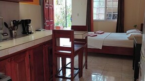 A compact hotel room with a kitchenette, a bed, a chair, and a window with curtains.