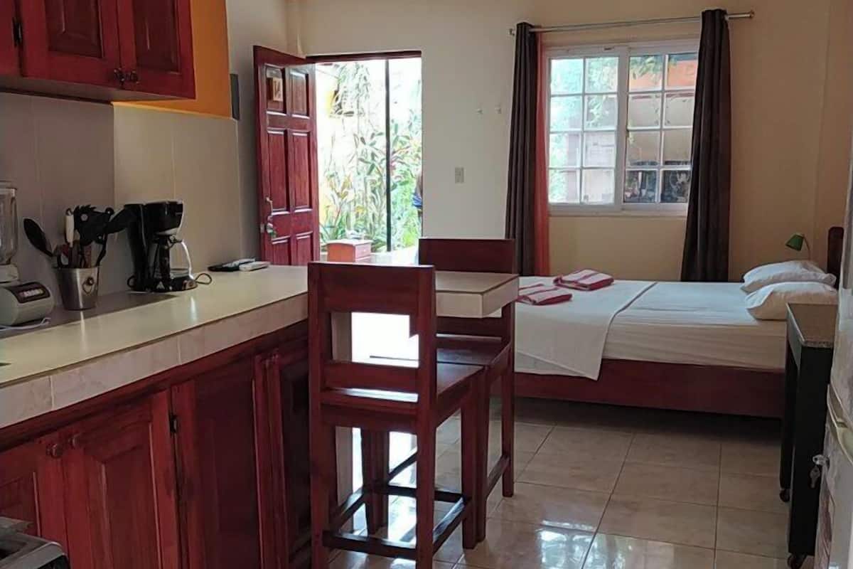 A compact hotel room with a kitchenette, a bed, a chair, and a window with curtains.