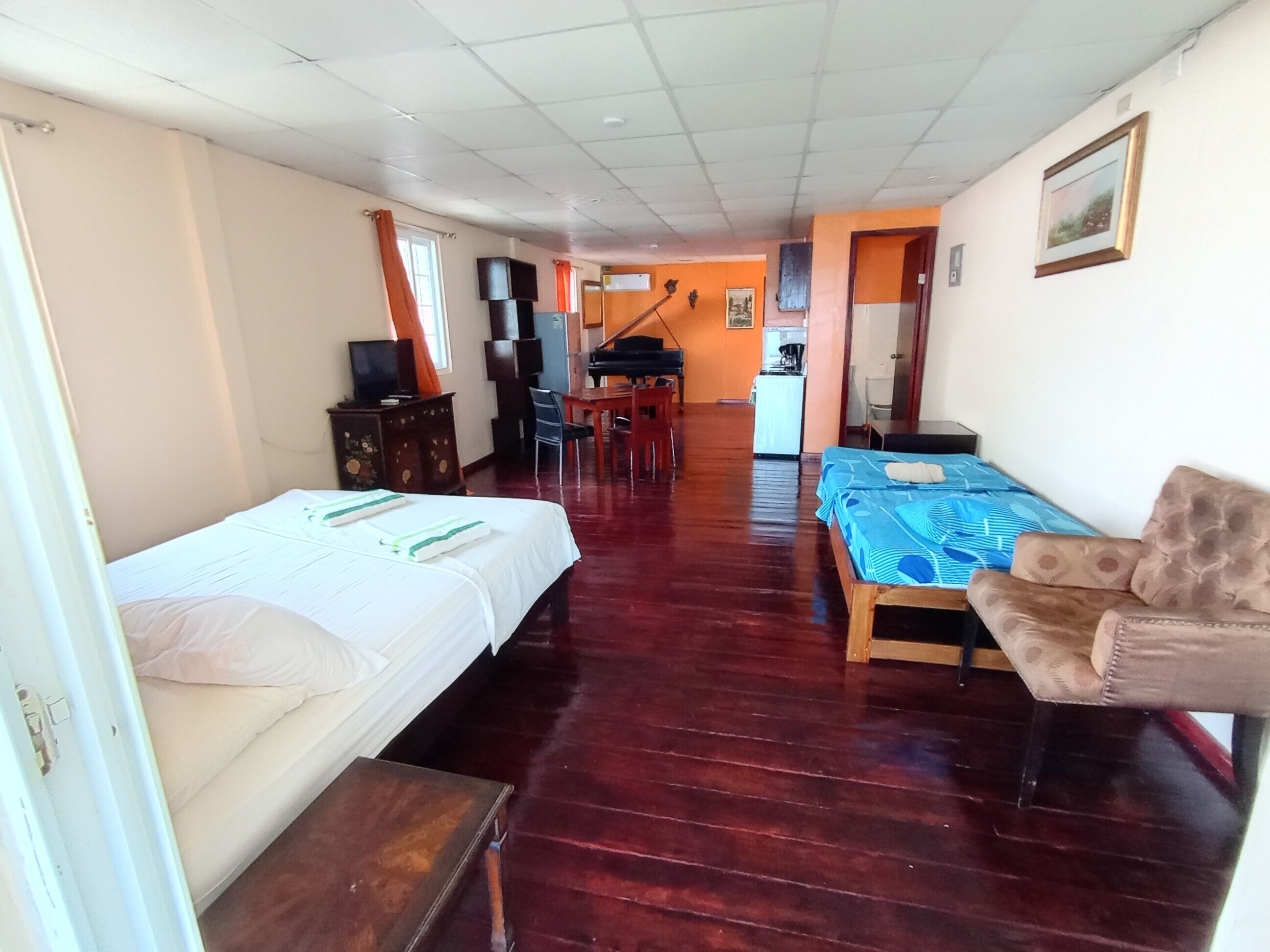 A room with a bed, a sofa, a desk, a chair, a piano, and a kitchenette.