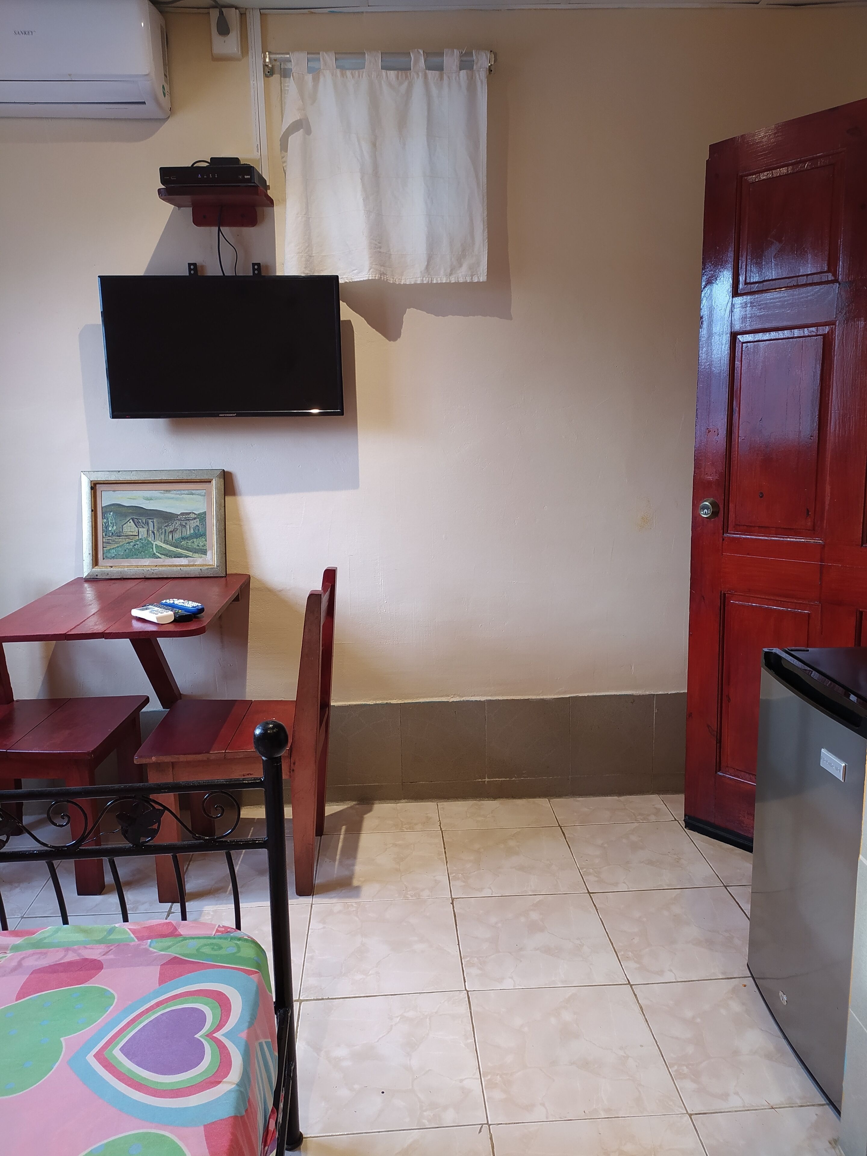 A small room with a bed, a table with chairs, a TV mounted on the wall, a door, and a refrigerator.
