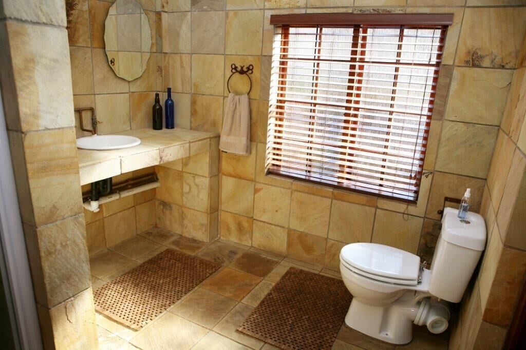 Een badkamer met een wit wastafelmeubel, spiegel en toilet, stenen tegels, en een raam met jaloezieën.