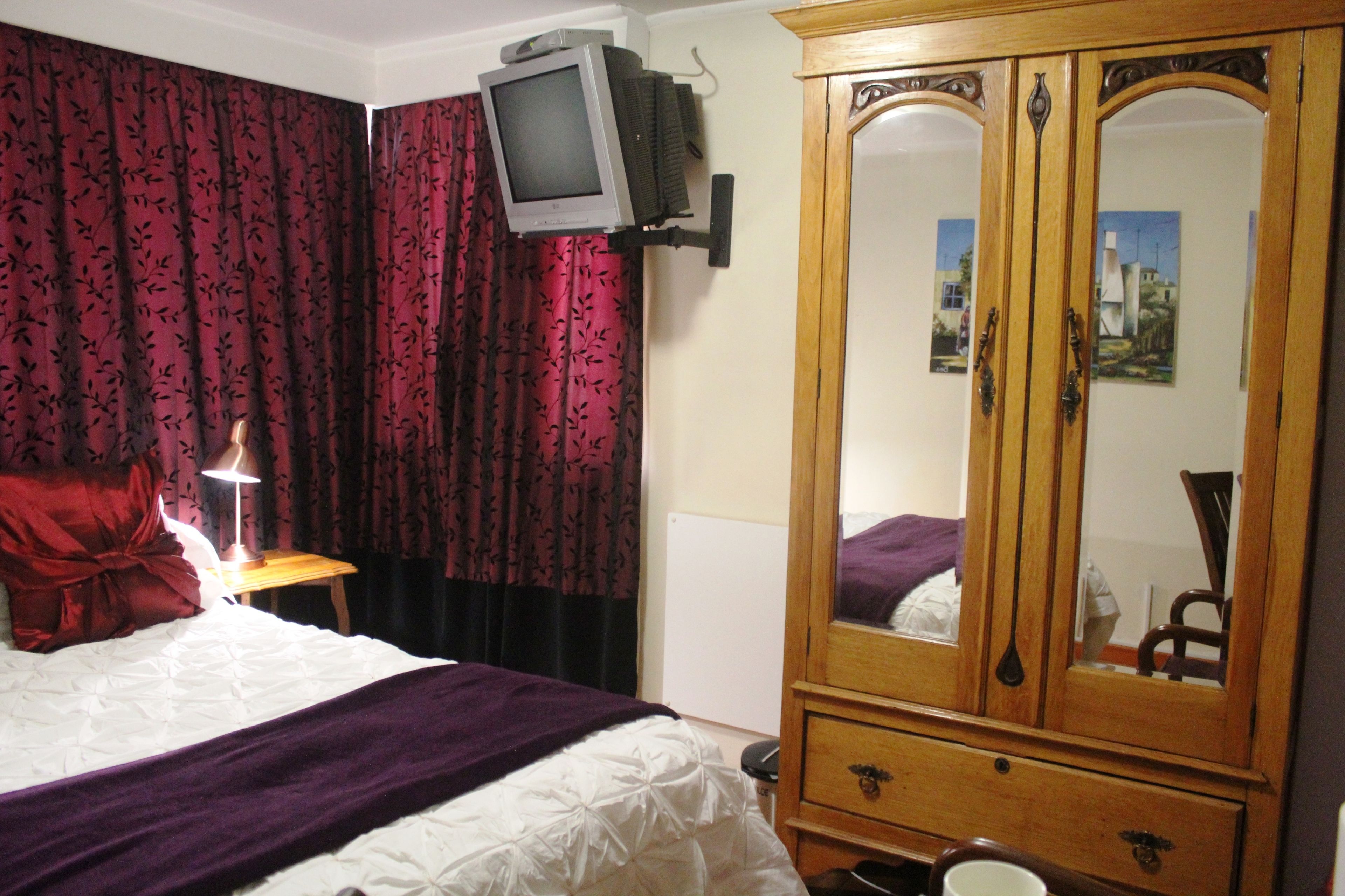 Quarto de hotel com uma cama, guarda-roupa de madeira, televisão na parede e janela com cortinas.