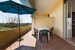 Uma varanda com mesa e cadeiras, um guarda-sol azul e vista para o campo.