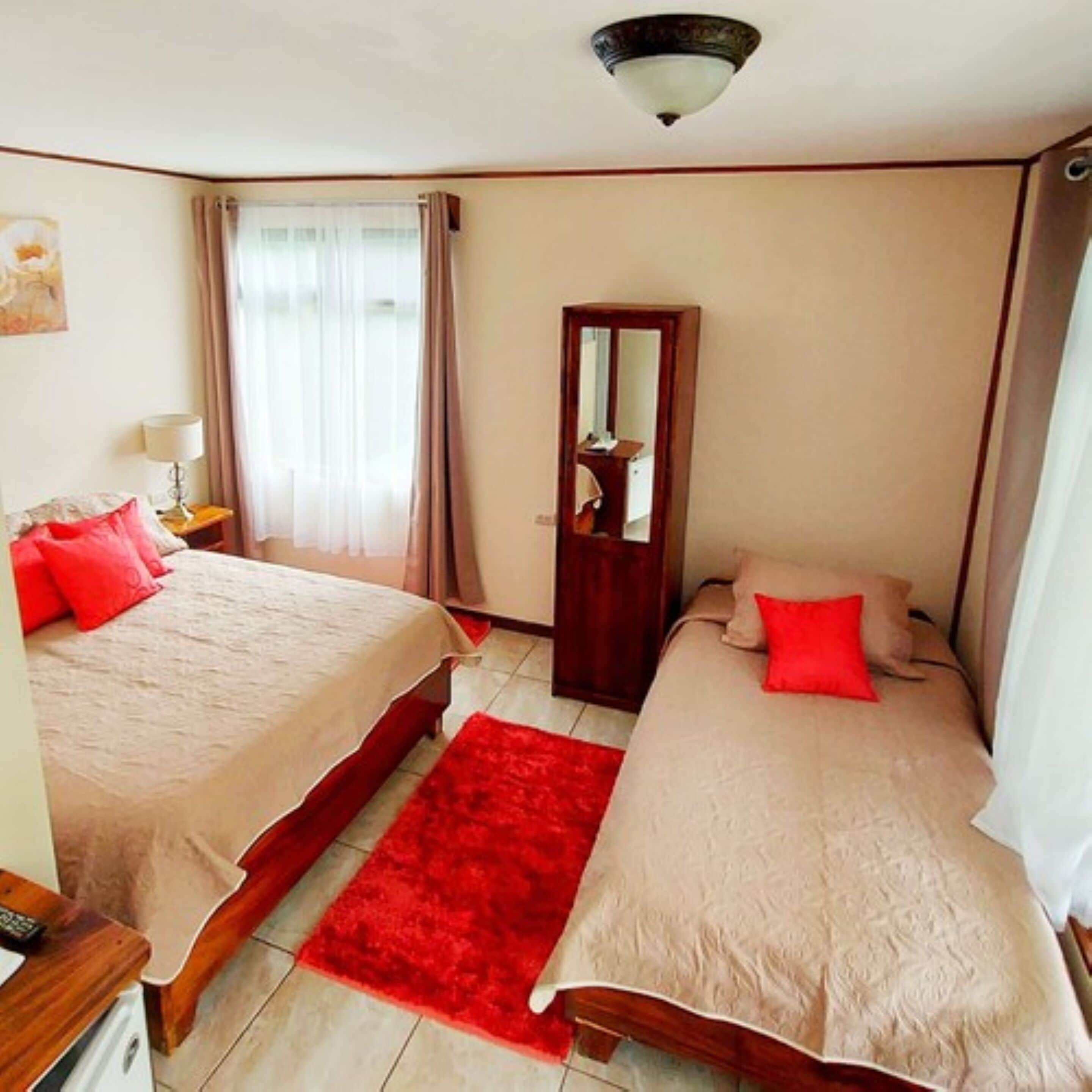 Un dormitorio con dos camas, un tapete rojo, una mesita de noche, una lámpara, un espejo y una puerta.