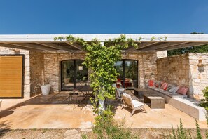 A modern outdoor patio with stone walls, a wooden pergola, and a seating area with cushions and a table. - La Gastalena (Ostuni)