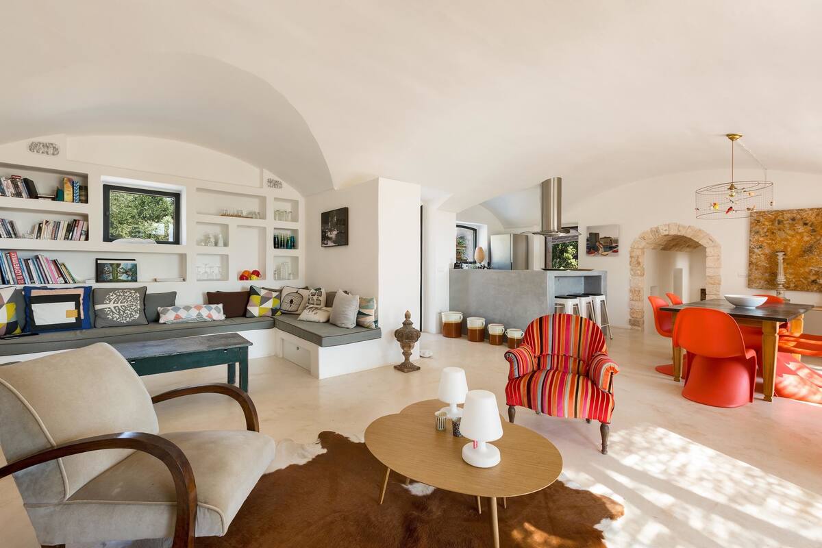 A spacious living area with a sofa, a coffee table, and a striped armchair. There is a bookshelf and a dining area with orange chairs.
