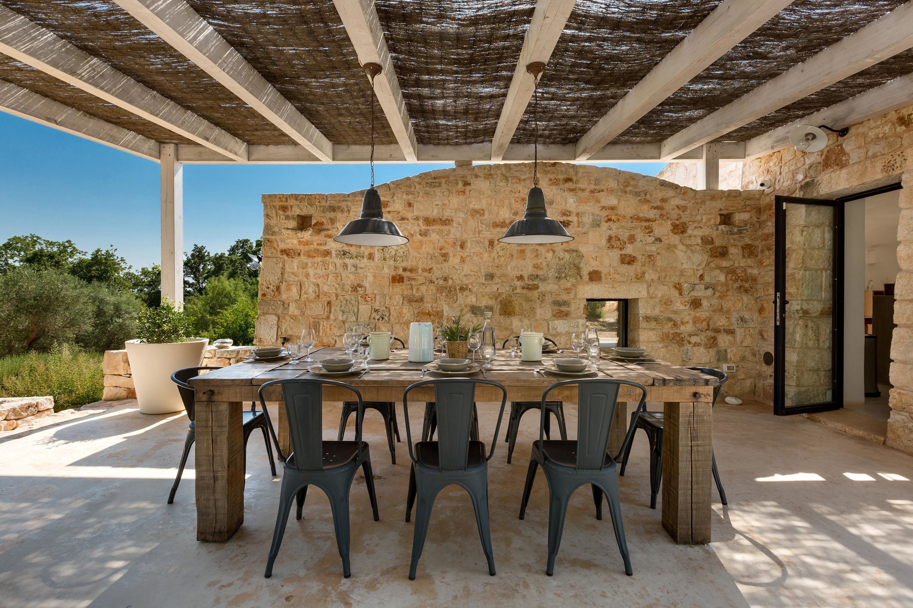 An outdoor dining area with a stone wall backdrop, a wooden table set for six, and two hanging pendant lights.