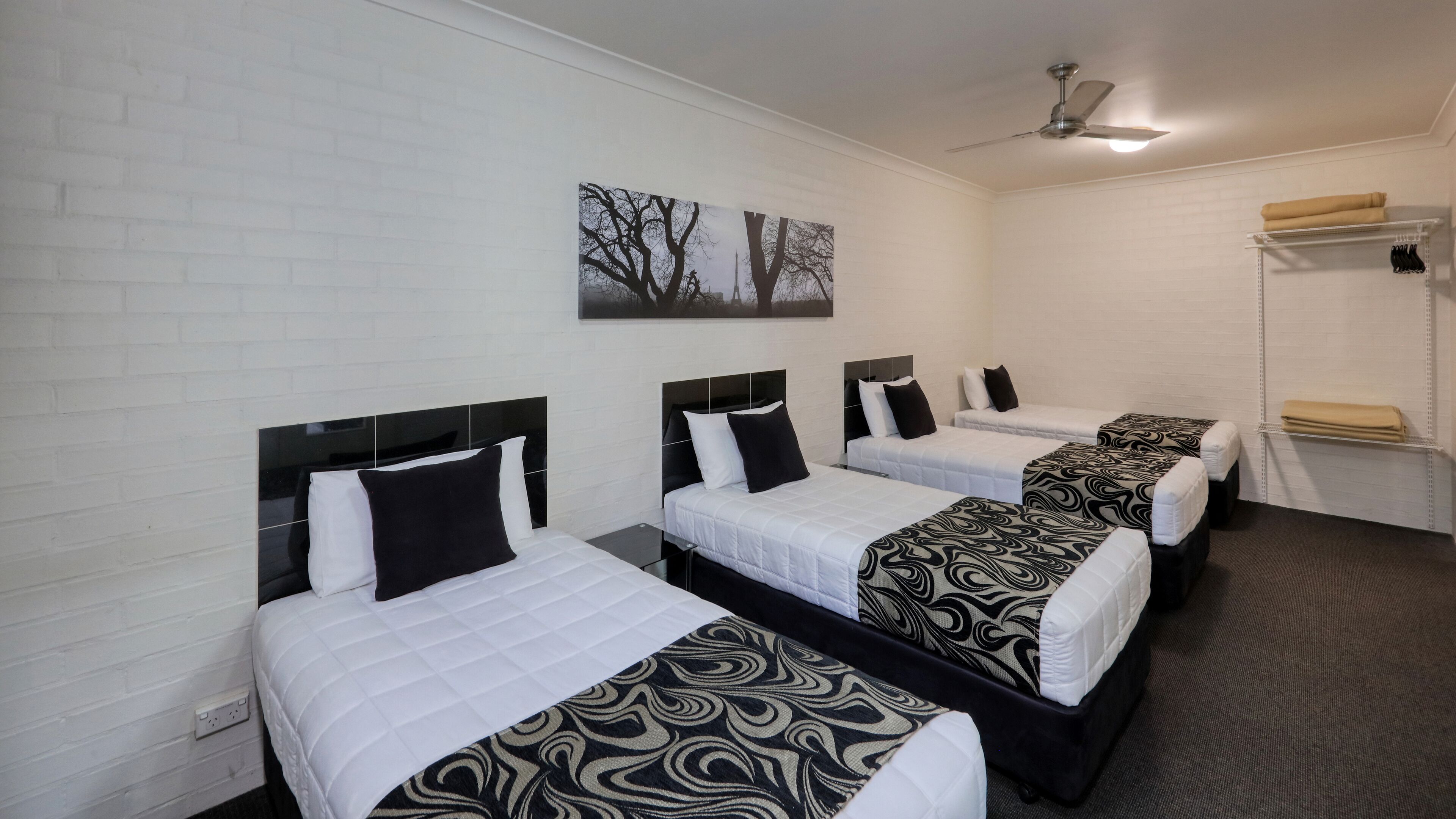 A hotel room with four single beds, each with a black and white patterned bedspread. There is a ceiling fan, a wall-mounted artwork depicting trees, and a white brick wall.