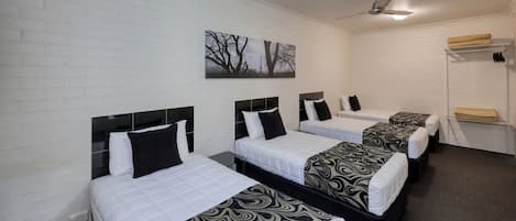 A hotel room with four single beds, each with a black and white patterned bedspread. There is a ceiling fan, a wall-mounted artwork depicting trees, and a white brick wall.