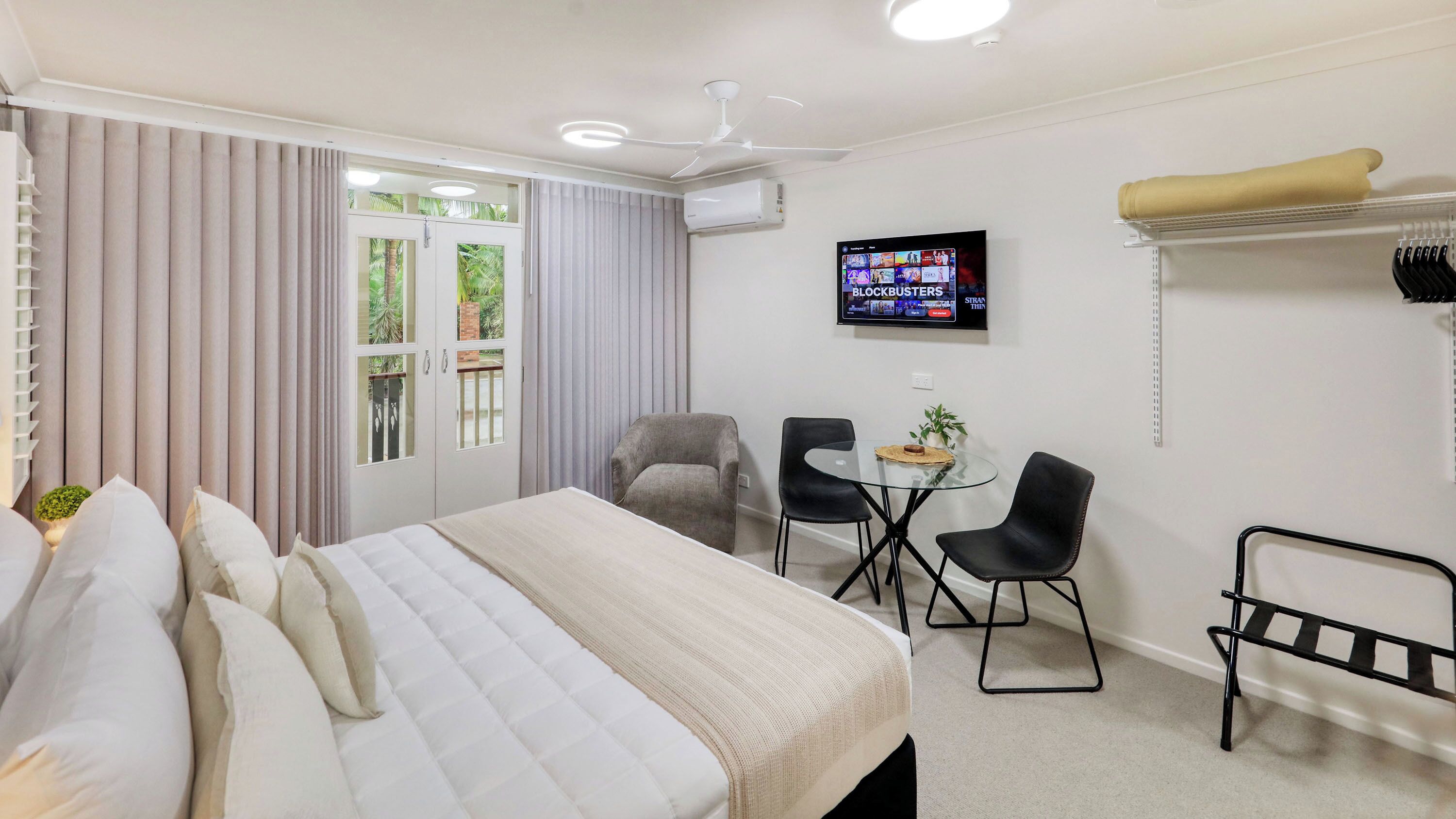Ein Hotelzimmer mit Bett, Fernseher, einem kleinen Tisch mit Stühlen und einem Balkon mit Vorhängen.