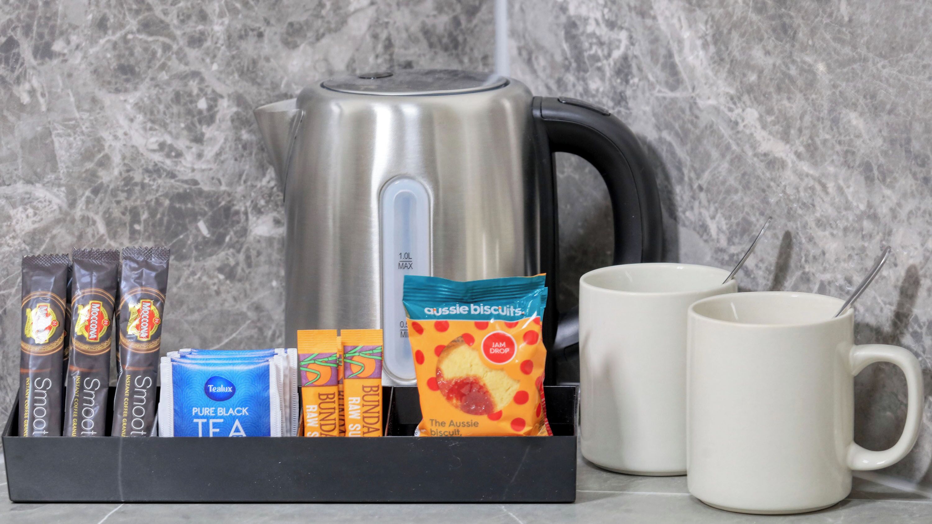 A kitchen setup with a kettle, instant coffee packets, a tea box, a packet of biscuits, and two white mugs.