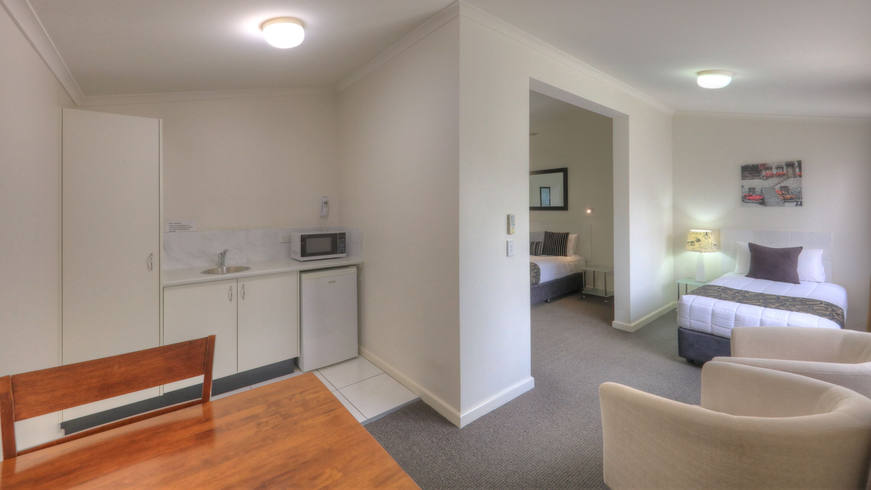 A compact hotel room with a kitchenette, a single bed, a bedside table with a lamp, and a chair.