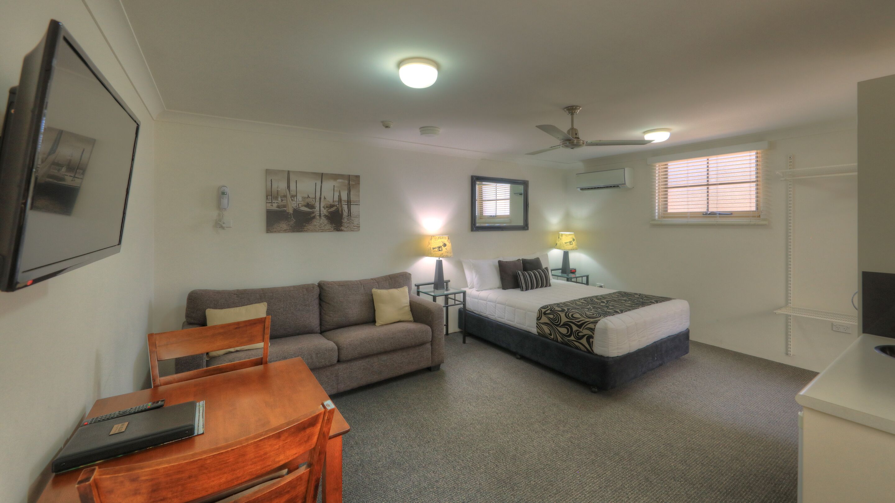 Habitación de hotel compacta con cama, sofá, escritorio y silla. Tiene dos lámparas de noche, ventilador de techo y aire acondicionado instalado en la pared.