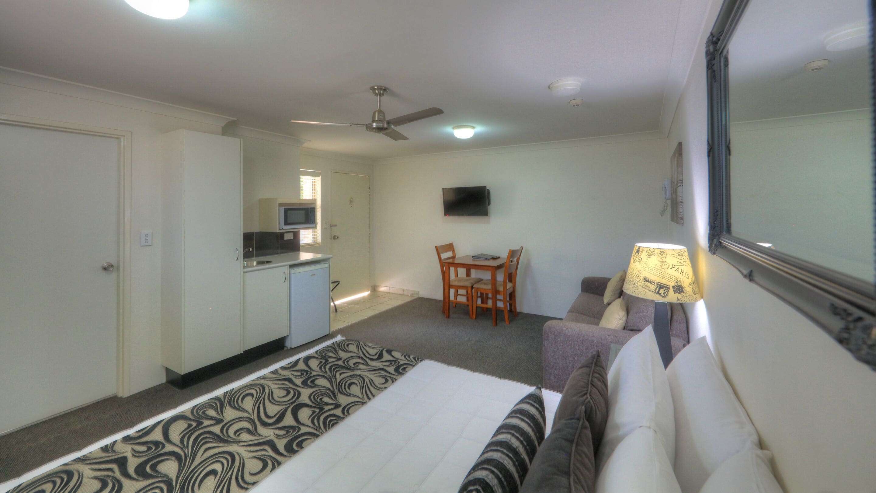 A hotel room with a bed, a desk with a chair, a television on the wall, and a ceiling fan.