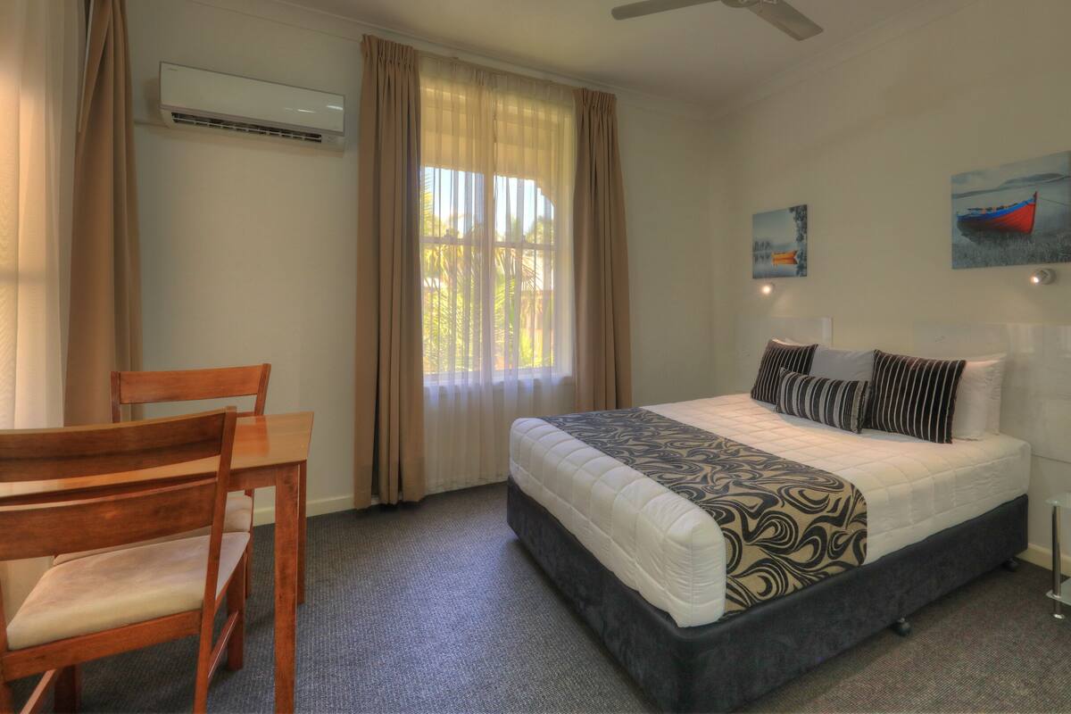 Un dormitorio con cama, una silla, un escritorio, aire acondicionado y dos cuadros en la pared.