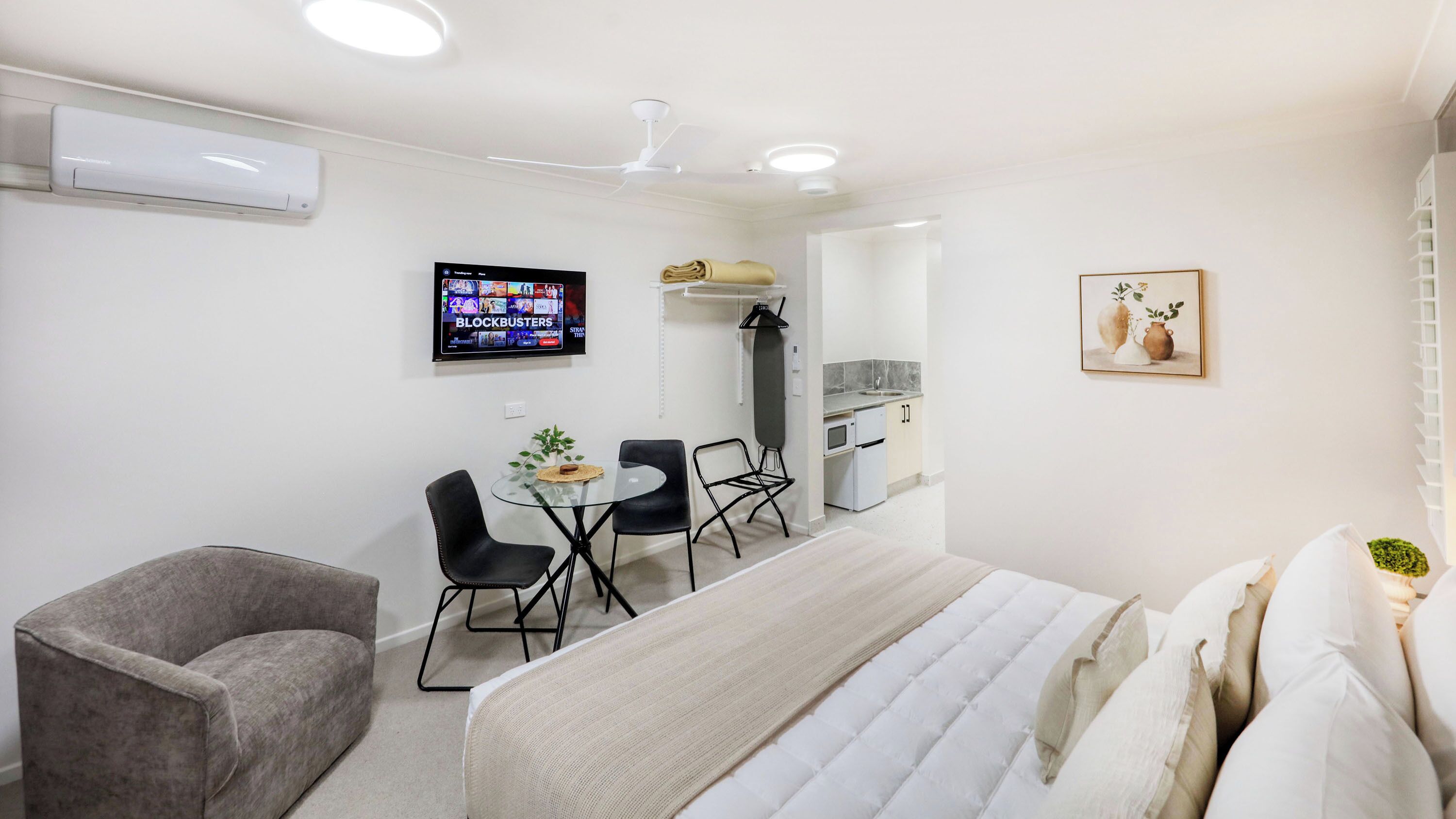 Habitación de hotel pequeña con cama, una mesita con sillas, televisión, kitchenette y un cuadro en la pared.