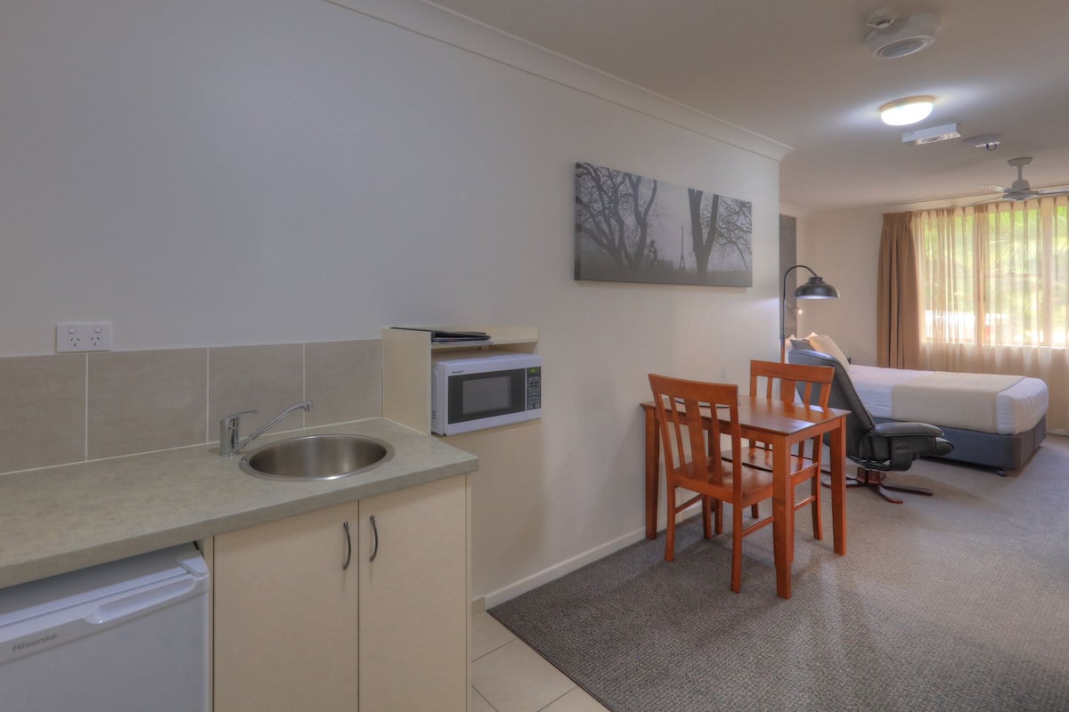 Cocina compacta con microondas integrado, lavaplatos y lavamanos. Zona de comedor con mesa y sillas, y cama con ventana.