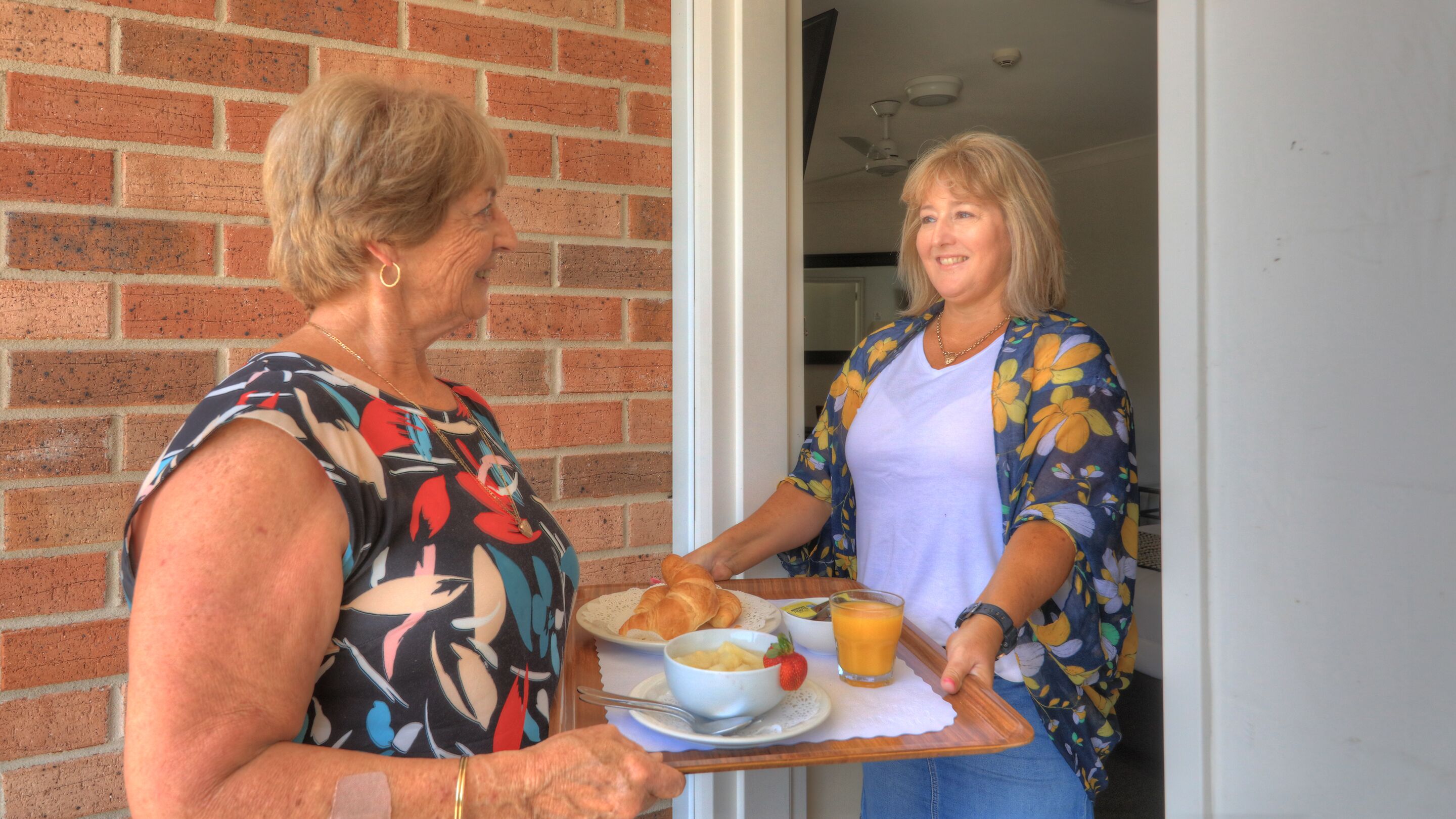 Twee vrouwen met een dienblad met croissants, een fruitschaal en een glas sinaasappelsap.