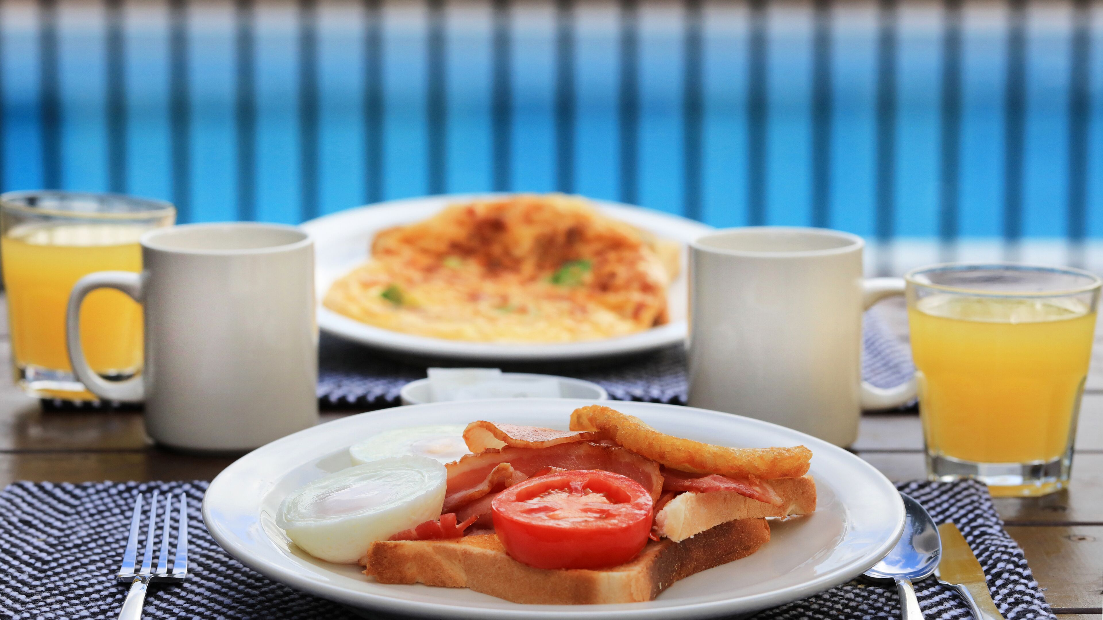 A breakfast setting with toast, eggs, bacon, and tomato on a plate, accompanied by two mugs and glasses of orange juice.