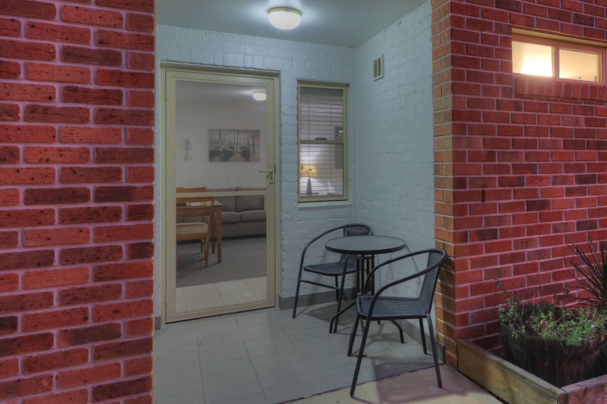 Un patio con pared de ladrillo, una pequeña mesa con sillas y una planta en maceta.