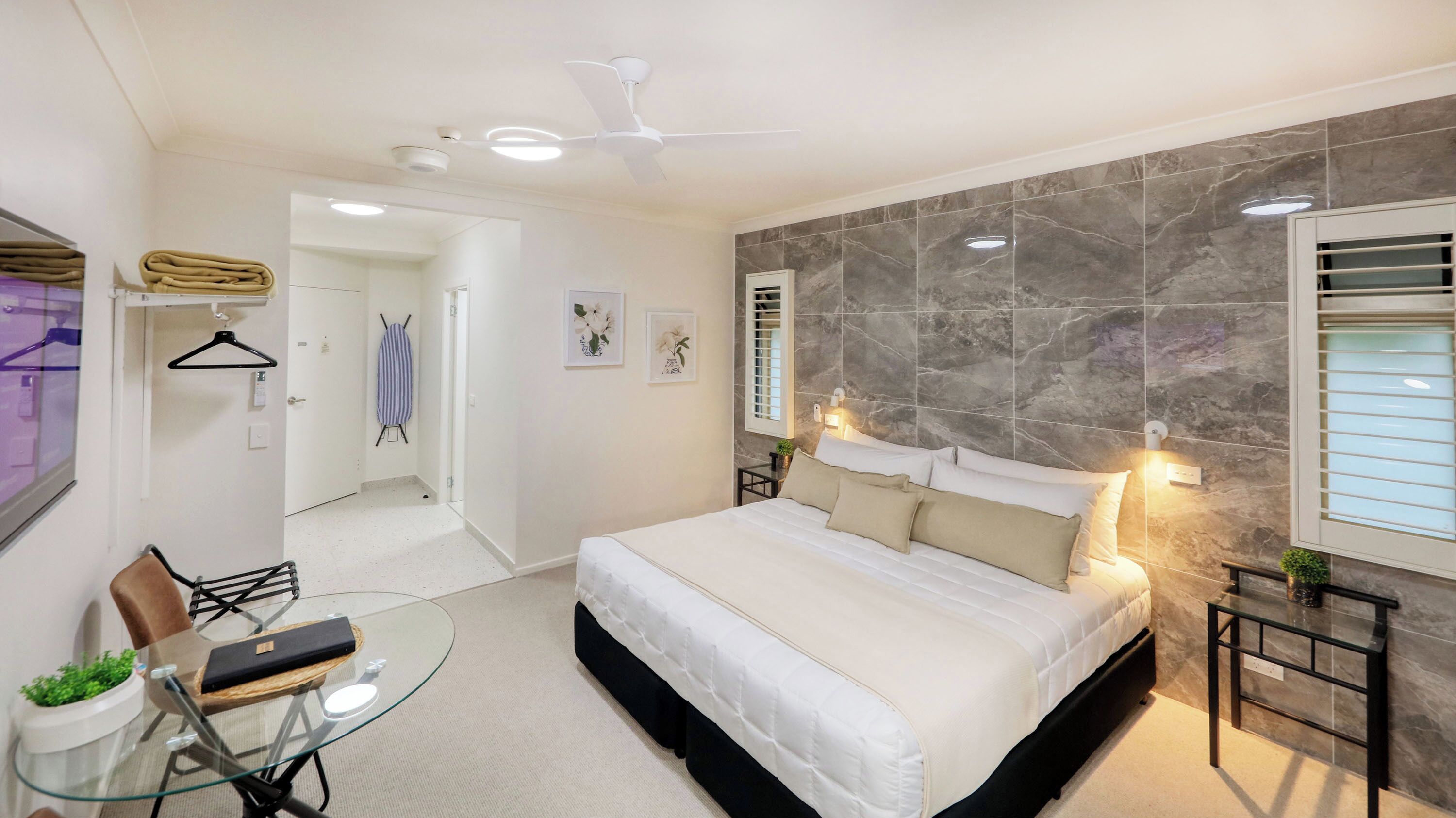 A modern bedroom with a large bed, a glass table, a hanging clothes rack, and a view of the hallway.