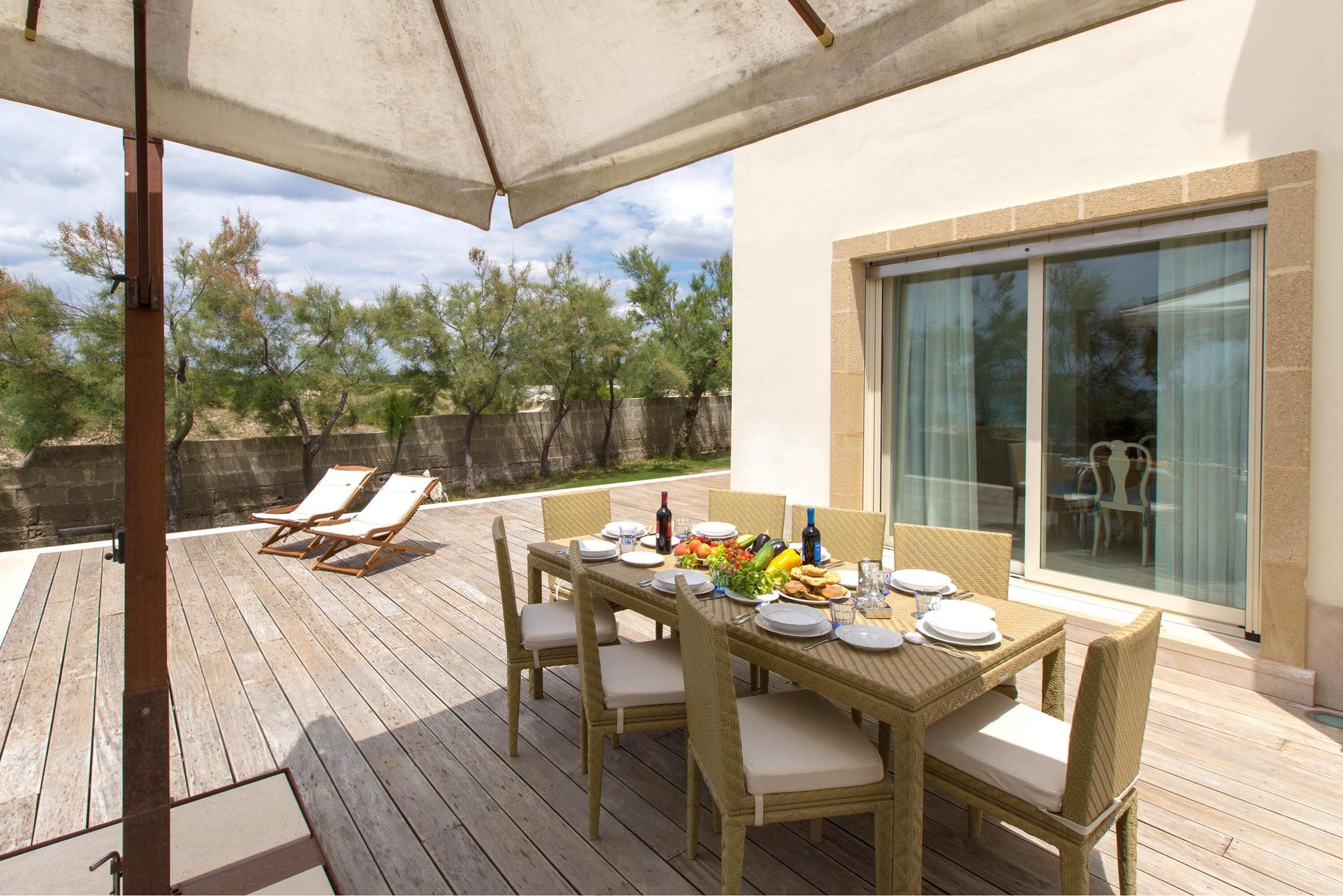 Une terrasse en bois avec une table dressée pour les repas, des chaises d’extérieur et un grand parasol pour faire de l’ombre.