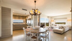 A spacious living area with a round wooden table, white chairs, a sofa, and a chandelier.