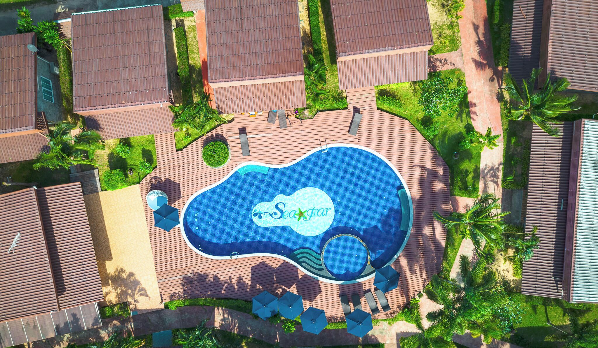 An aerial view of a resort with a central swimming pool, surrounded by wooden deck areas and lush greenery.