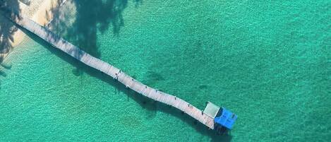 Une vue aérienne d’une longue jetée en bois s’étendant dans une mer turquoise et limpide, avec une petite construction à son extrémité.