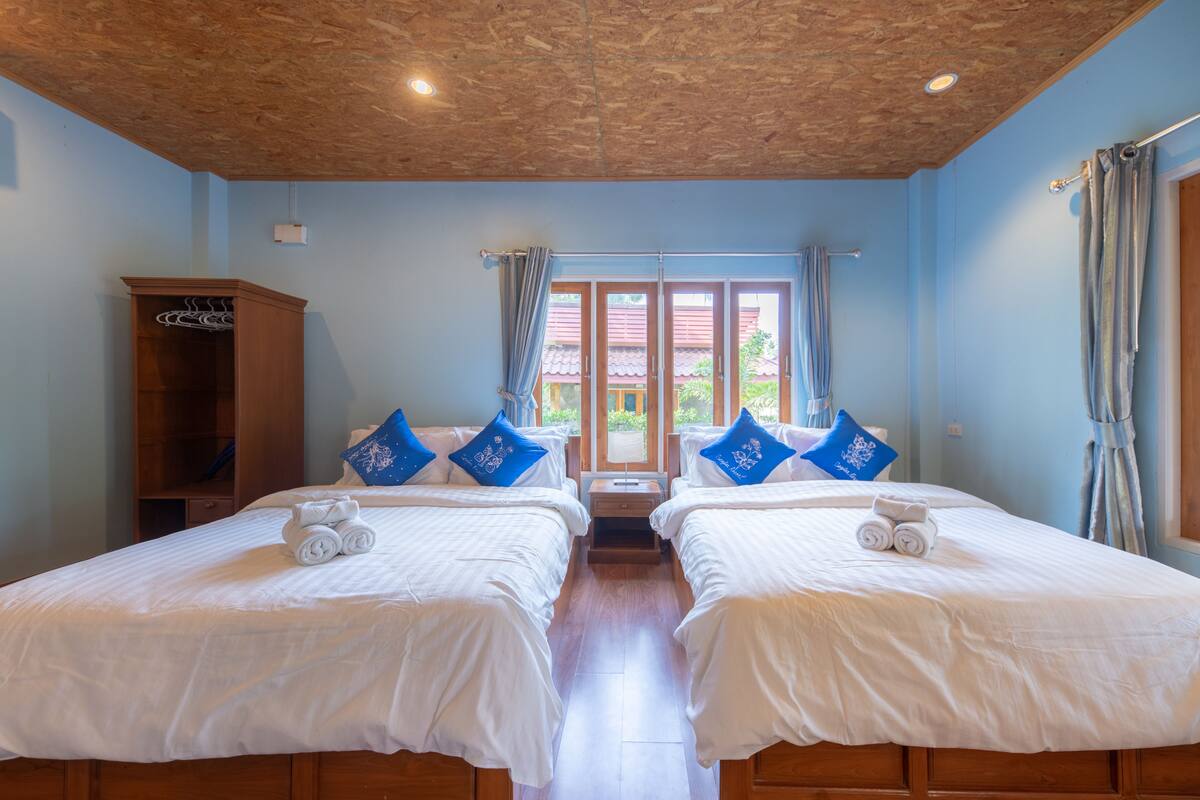 A hotel room with two beds, wooden headboard, a wardrobe, and a view of the outside through a window with curtains.