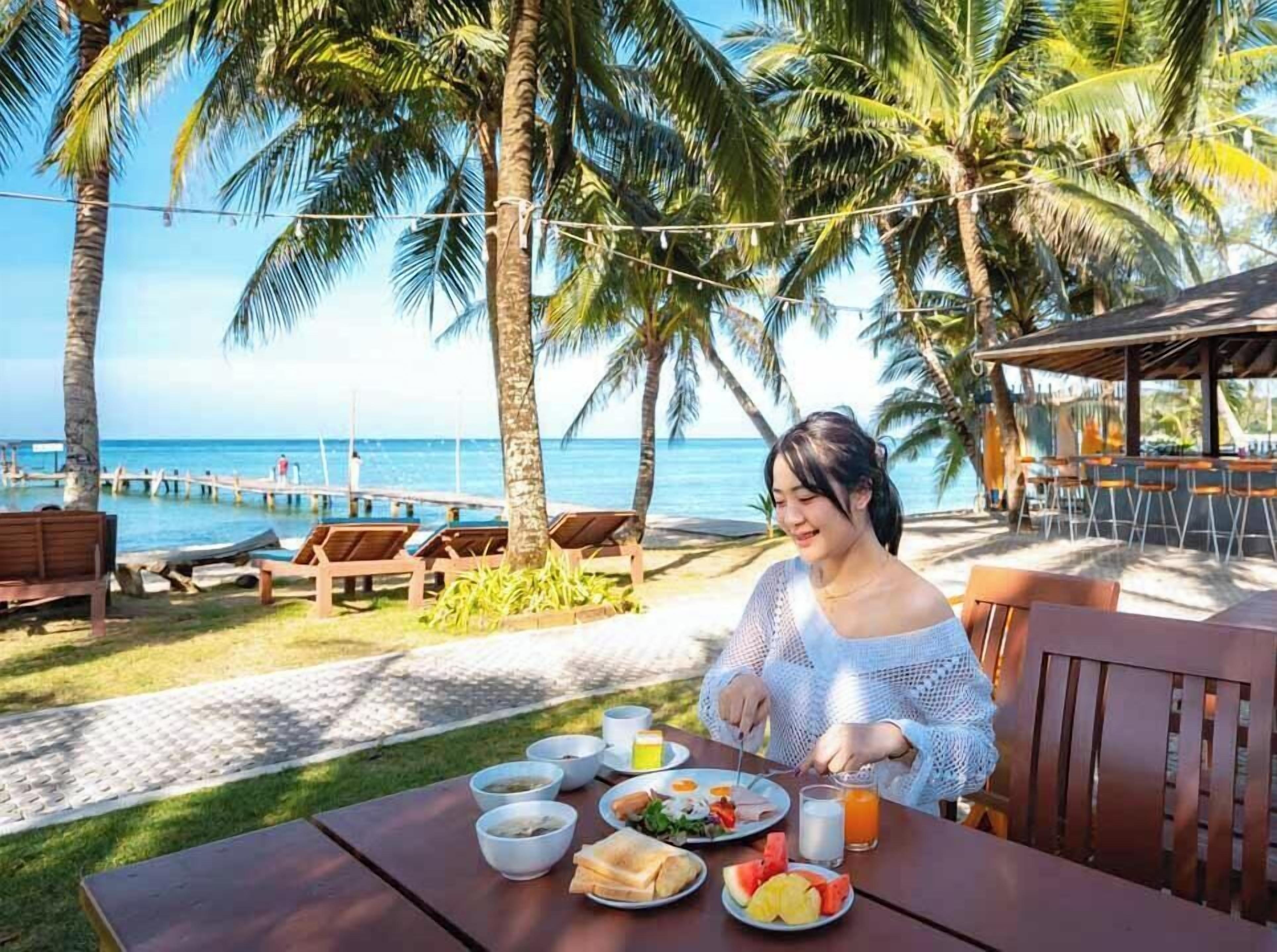 A dining area with a table set for two, overlooking a beach with palm trees and a pier.
