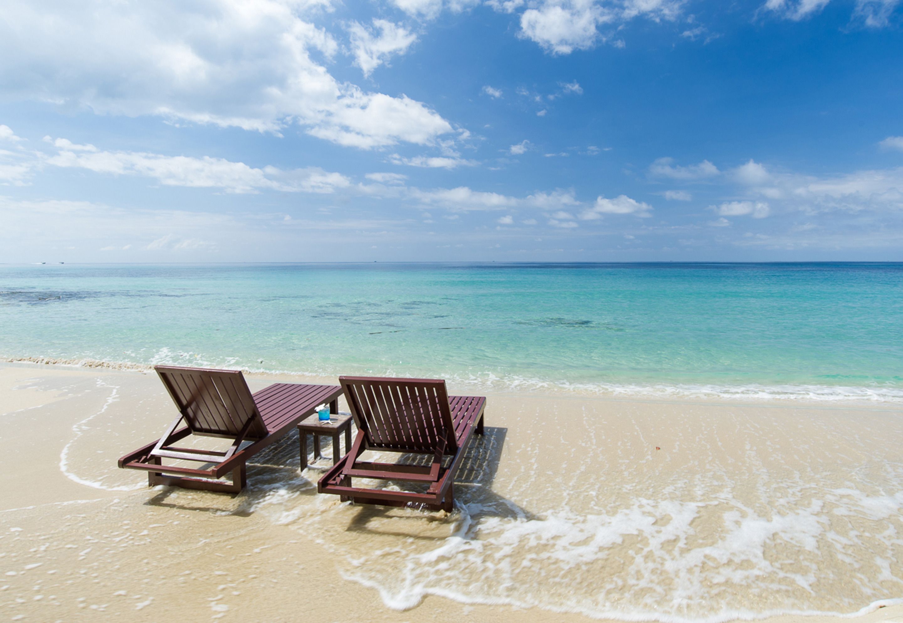 Two wooden lounge chairs on a sandy beach with clear turquoise water and a blue sky.