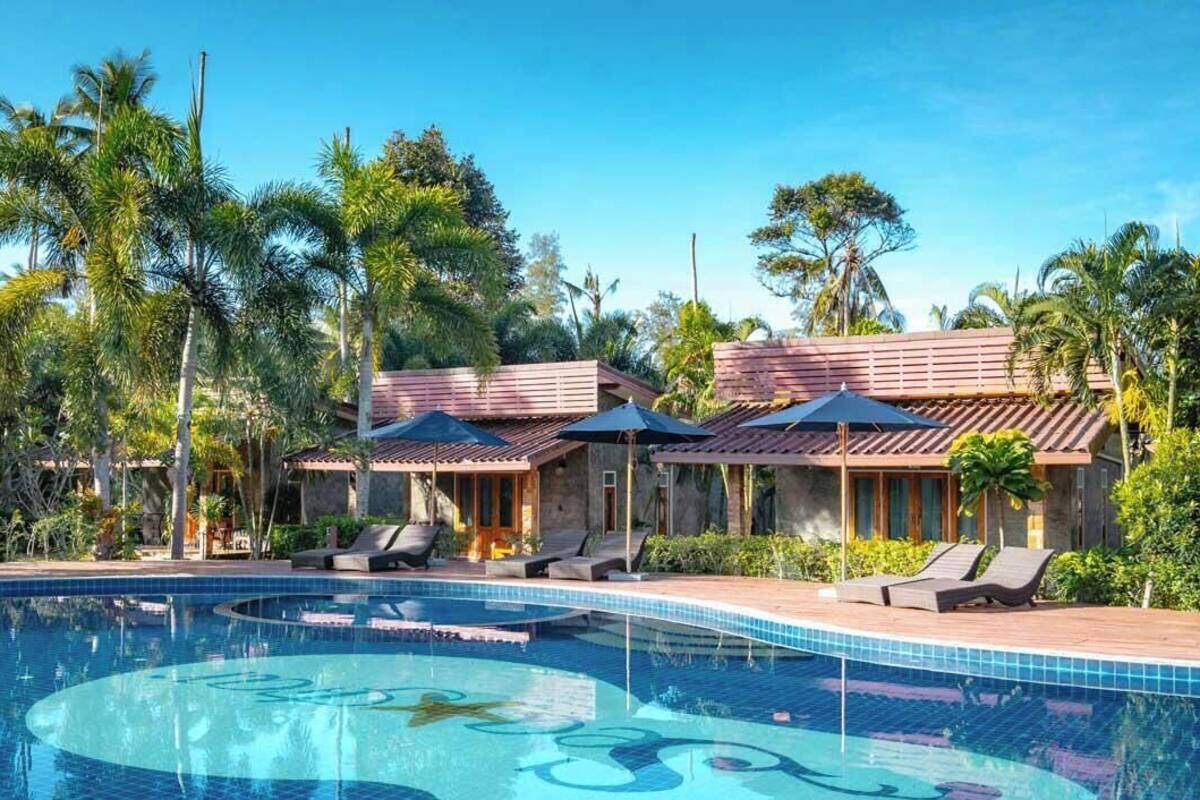 A swimming pool with lounge chairs and umbrellas, surrounded by palm trees and a wooden fence.