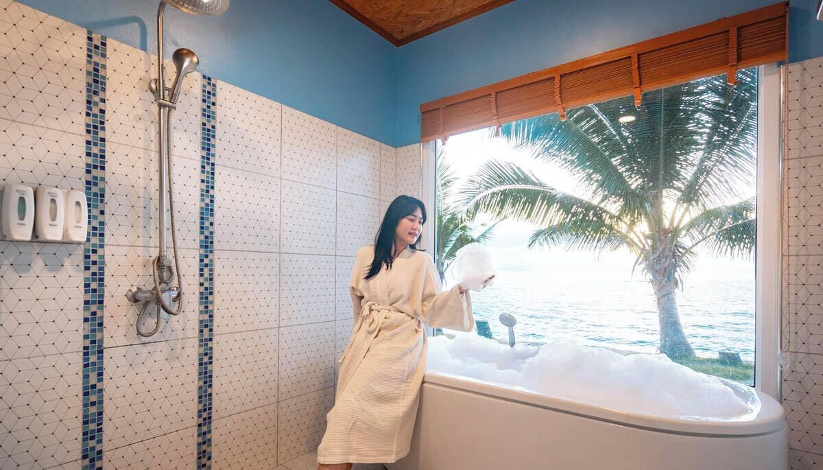 A bathroom with a bathtub, a shower, and a window with a view of palm trees and water.