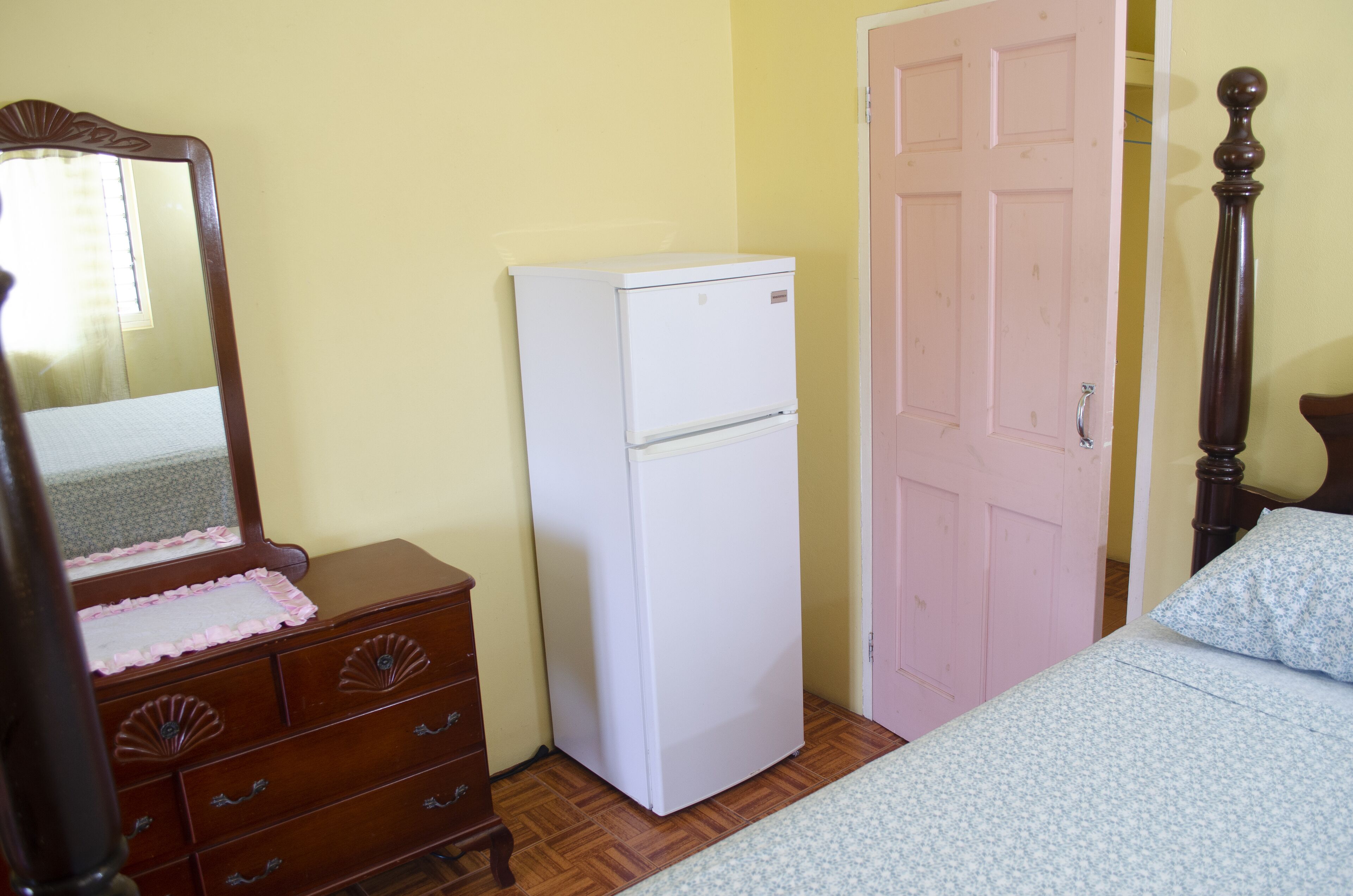 Une chambre avec une commode en bois, un réfrigérateur blanc, une porte rose et un lit recouvert d’une couvre-lit à motifs floraux.