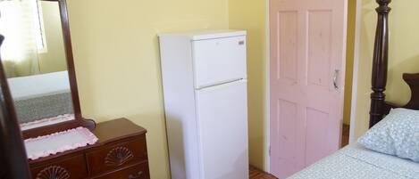 Une chambre avec une commode en bois, un réfrigérateur blanc, une porte rose et un lit recouvert d’une couvre-lit à motifs floraux.