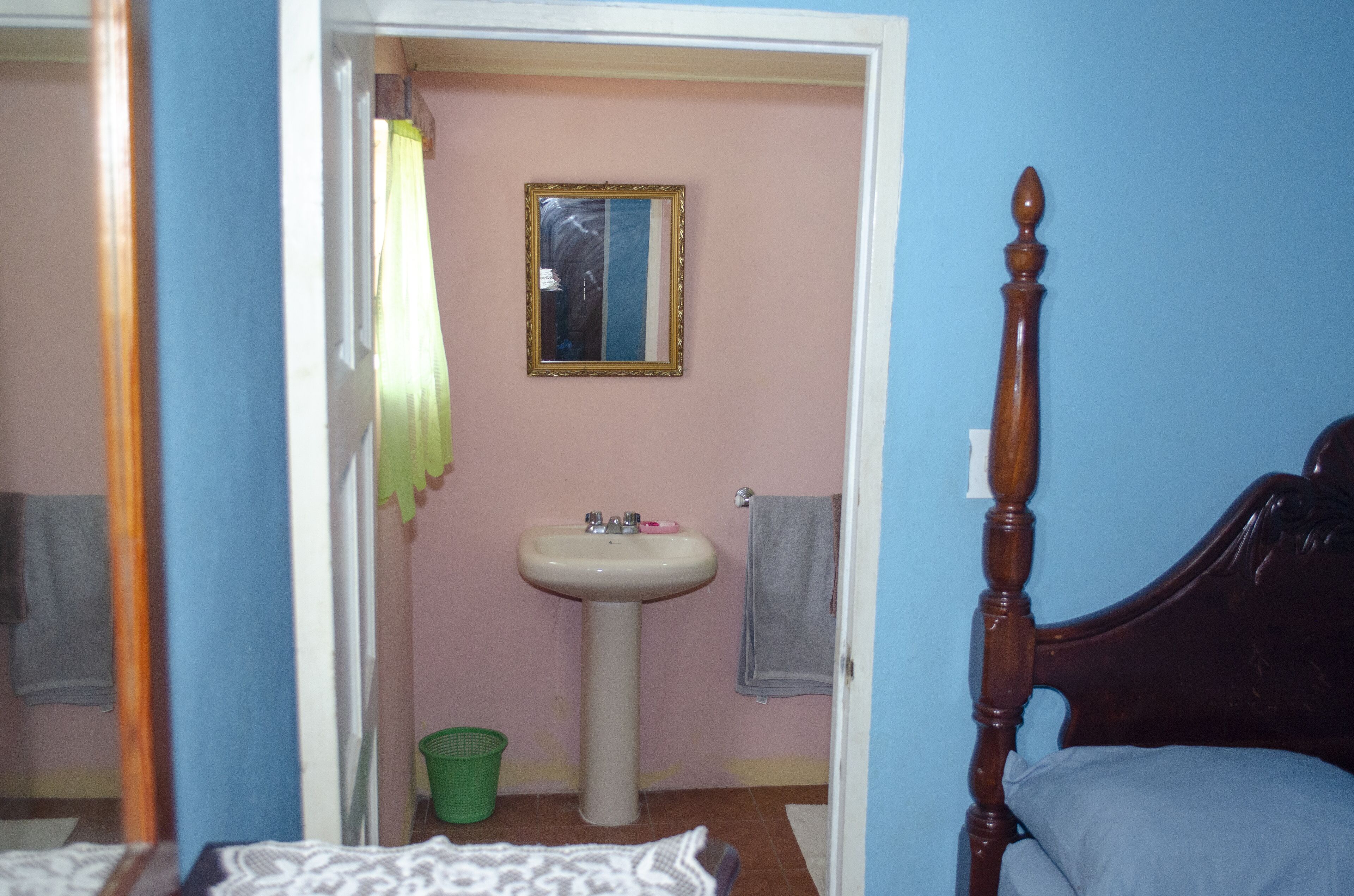 A bedroom with a wooden bed, a bedside table, a green trash bin, a white sink, and a mirror on the wall.