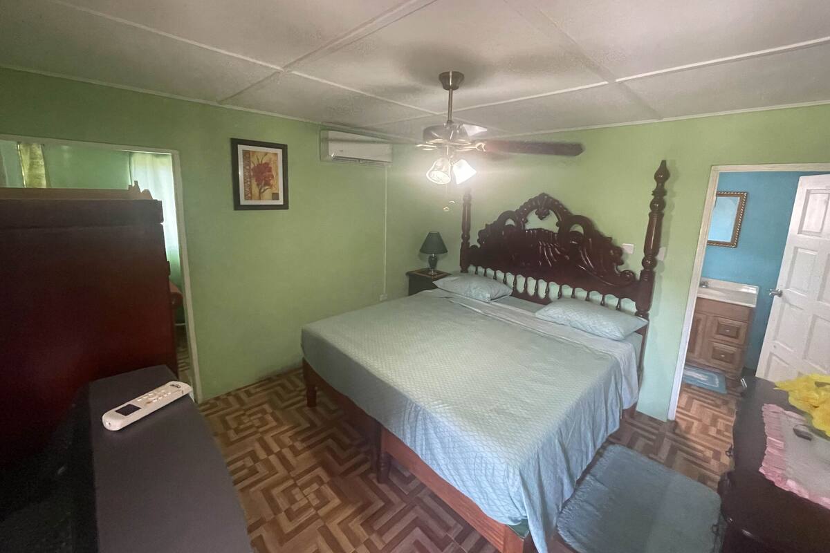 Une chambre à coucher comprenant un lit, un ventilateur de plafond, une table de chevet avec une lampe et une porte donnant sur une salle de bain.