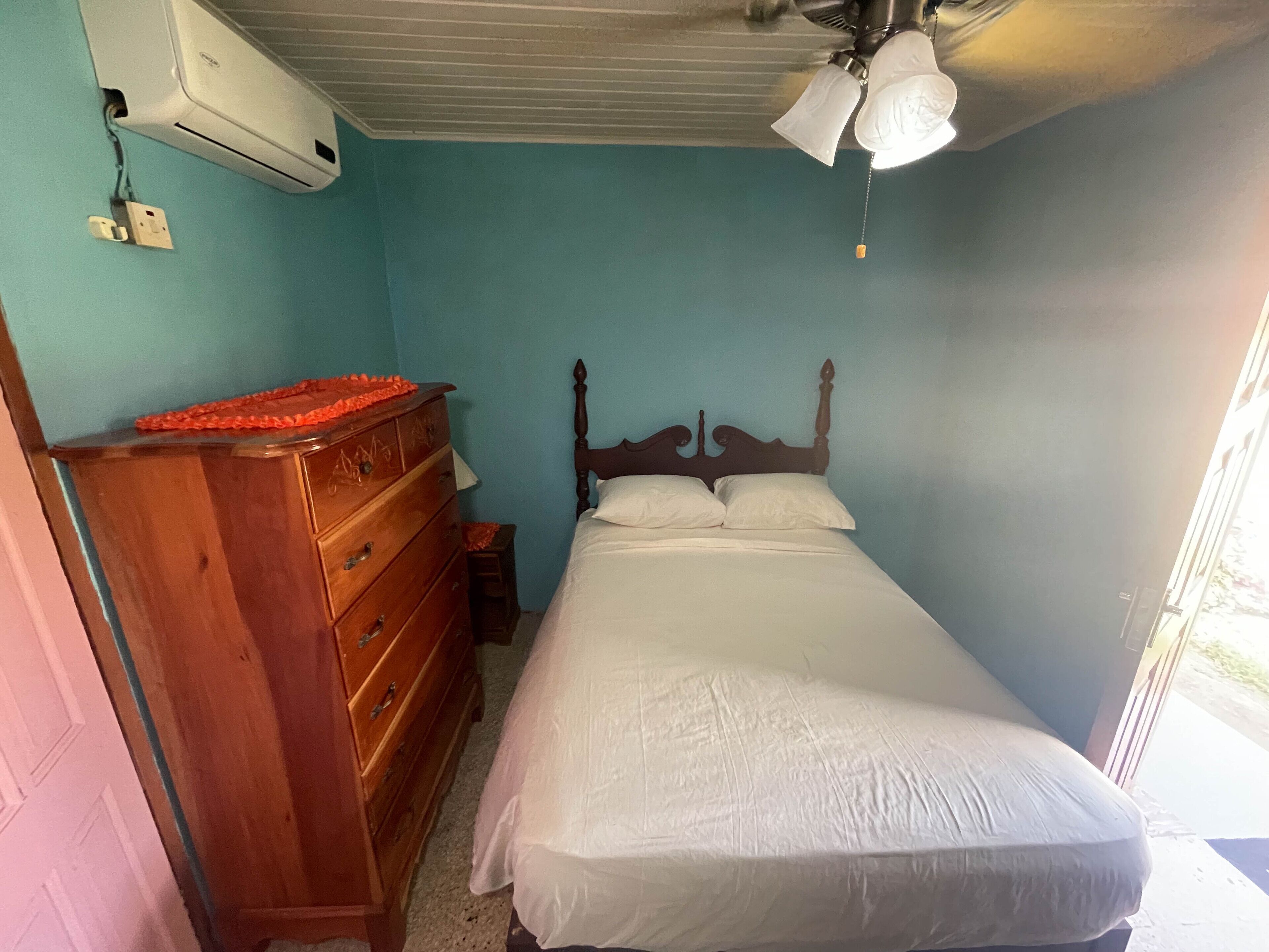 Een slaapkamer met een houten dressoir, een bed met witte beddengoed, een plafondventilator en een airconditioning.