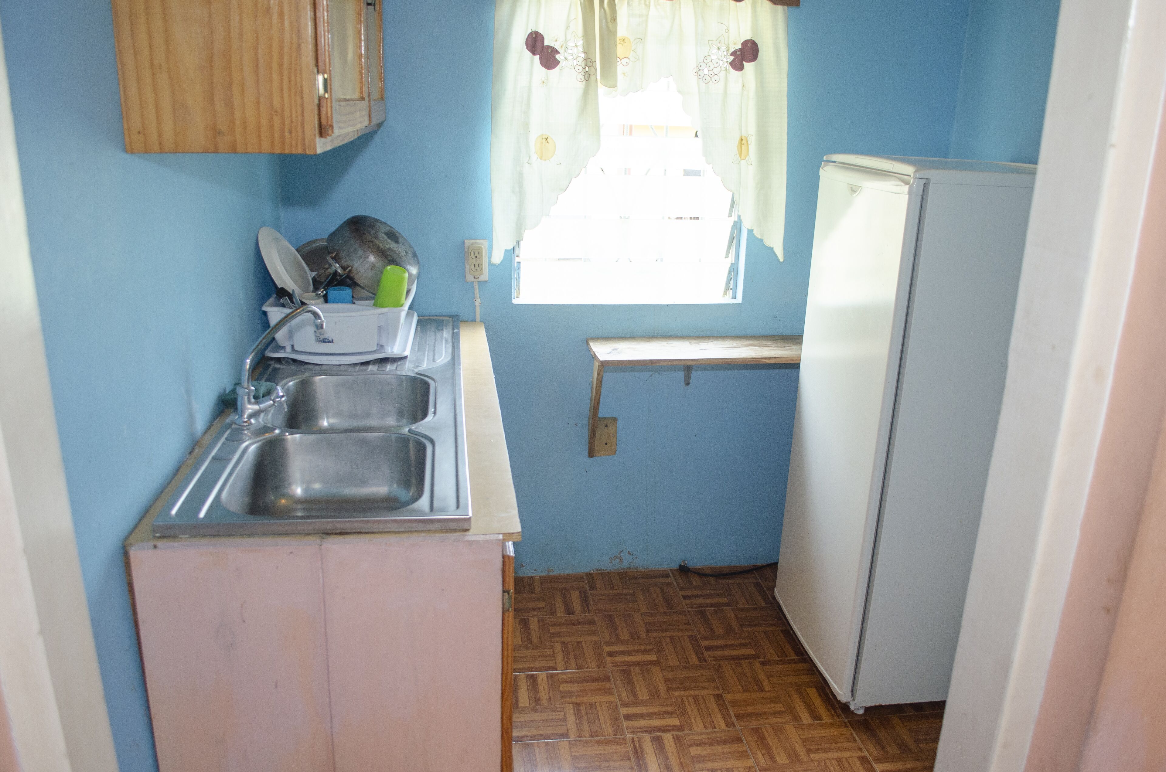 A compact kitchen with a sink, wooden cabinets, a small window with curtains, and a white refrigerator.