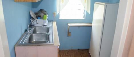 A compact kitchen with a sink, wooden cabinets, a small window with curtains, and a white refrigerator.